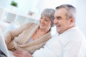 Picture of an eldery couple sitting next to each other smiling while they look down at an open laptop.