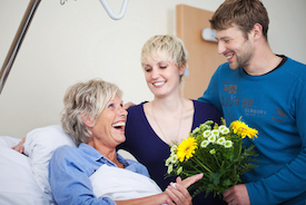 Picture of a female patient sitting up in a swing bed smiling as she receives a bouquet of pretty flowers from her loved ones (young man and young woman) who are smiling at her.