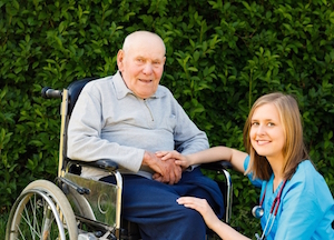 Picture of a female Nurse knelt down in front of a wheelchair that an elderly man is sitting in. They are both smiling at the camera.