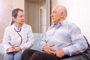 Picture of a female Physician that is holding a clipboard and pen and sitting next to an eldery man that is holding the side of his stomach.