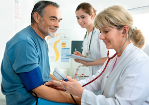 Picture of a male patient sitting on an exam table getting his blood pressure checked by a female Physician. There is a female Nurse in the background holding a clipboard and pen. They are all smiling.