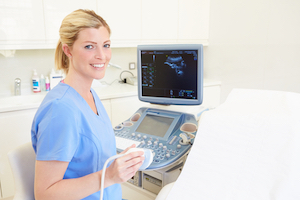 Picture of a female Ultrasound Tech standing next to an ultrasound/sonogram machine in an ultrasound room.