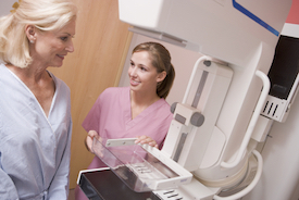 Picture of a female Nurse standing next to a mammogram machine looking at an older female patient who has a robe on.