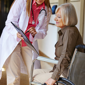 Picture of a female Physician bent over holding a pen that she is pointing on top of a clipboard as she shows a female patient that is in a wheelchair.