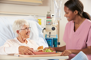 Picture of a female Nurse giving an elderly female patient her meal on a tray over her swing bed. They are smiling at each other.