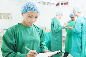 Picture of a surgeon and her surgeon team in the background in an Operation room. She is holding a clipnoard and pen about to write and her team is talking in the background.